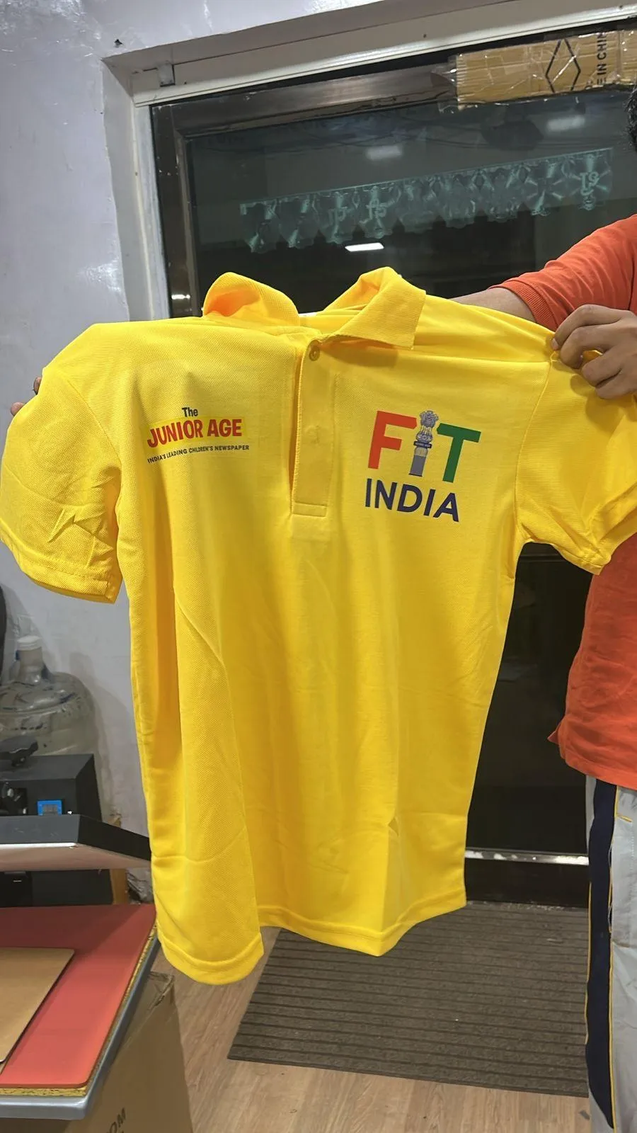 Yellow polo printed with “The Junior Age” and “Fit India” logos